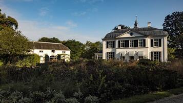 Een groot wit gebouw met meerdere ramen en een schoorsteen, omgeven door weelderige begroeiing en een grindpad.