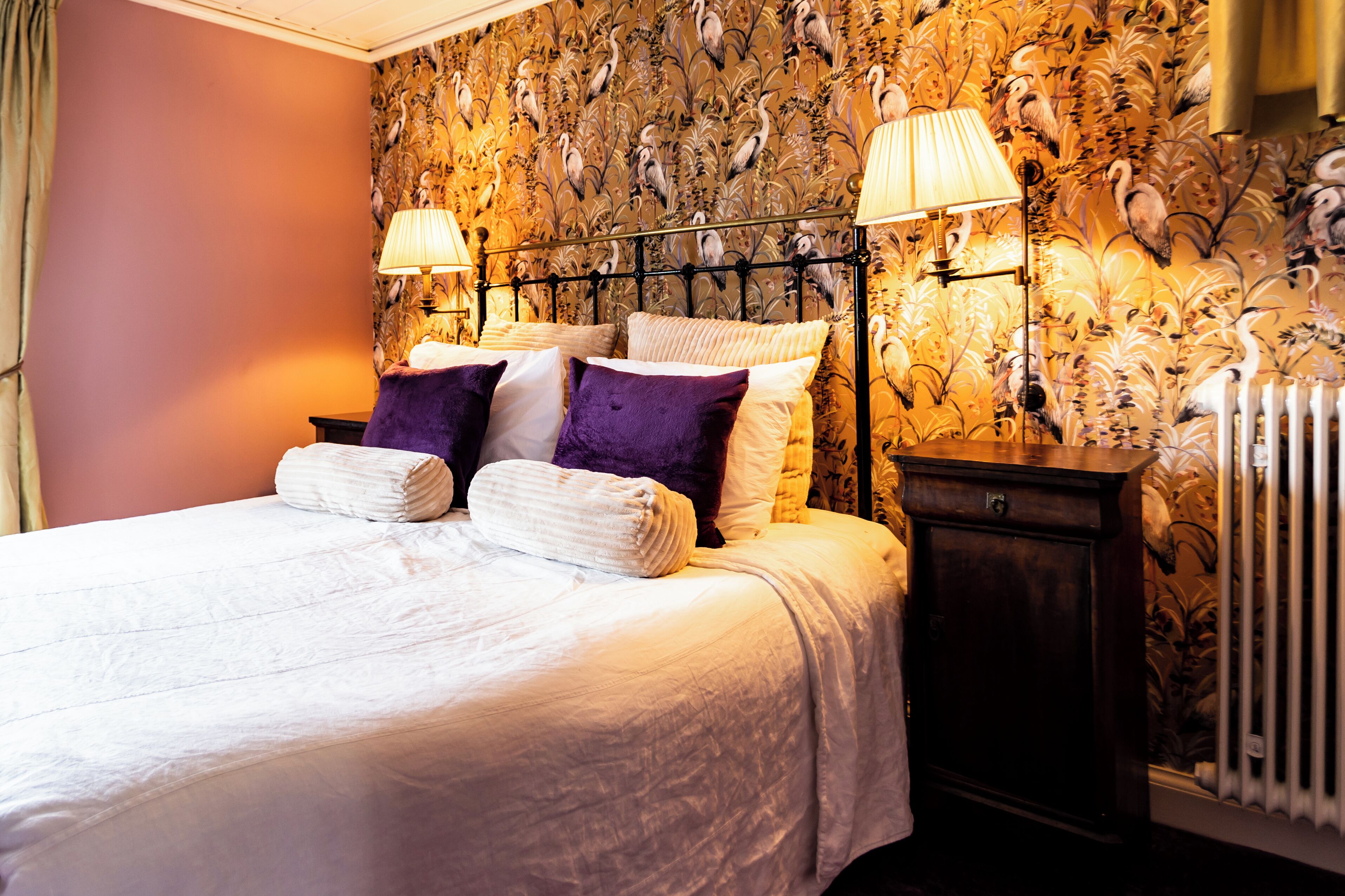 Cama bem arrumada com roupa de cama branca e roxa, duas luminárias de cabeceira e papel de parede floral.