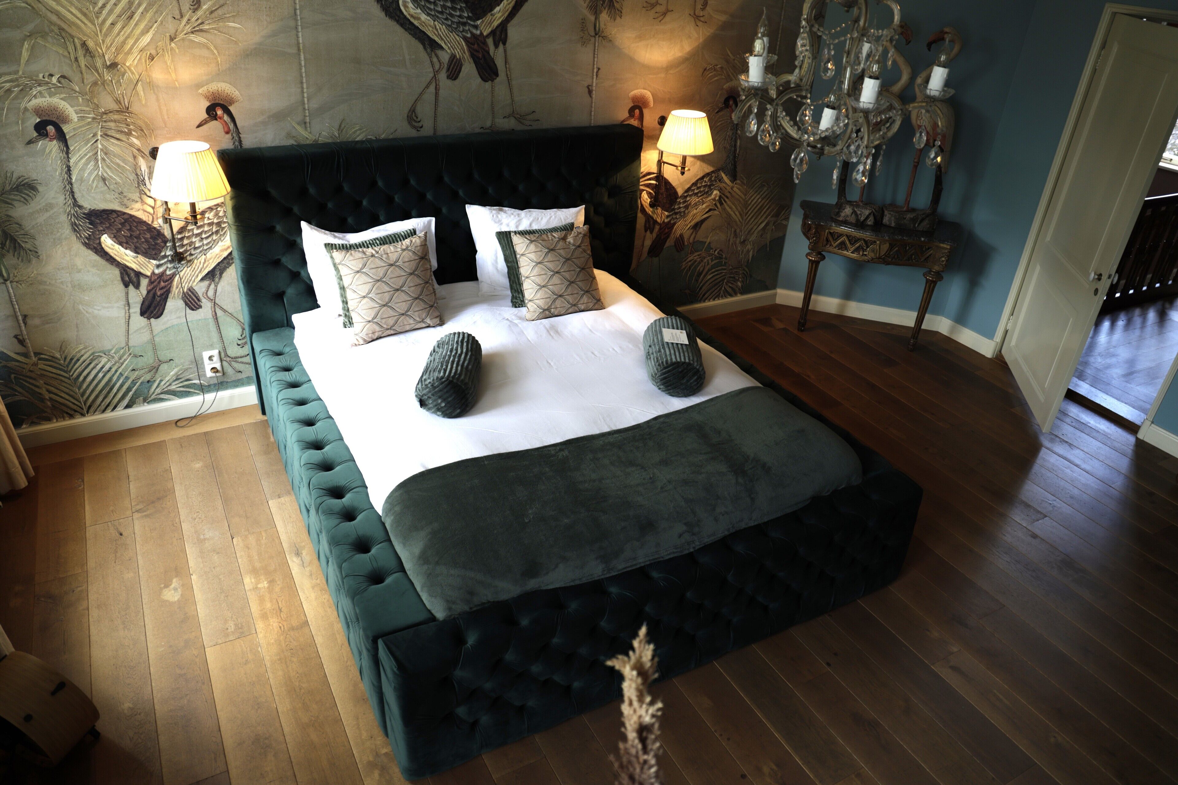 Ein Schlafzimmer mit einem großen Bett, einem dunkel gepolsterten Kopfteil und einem Wandbild von Vögeln.