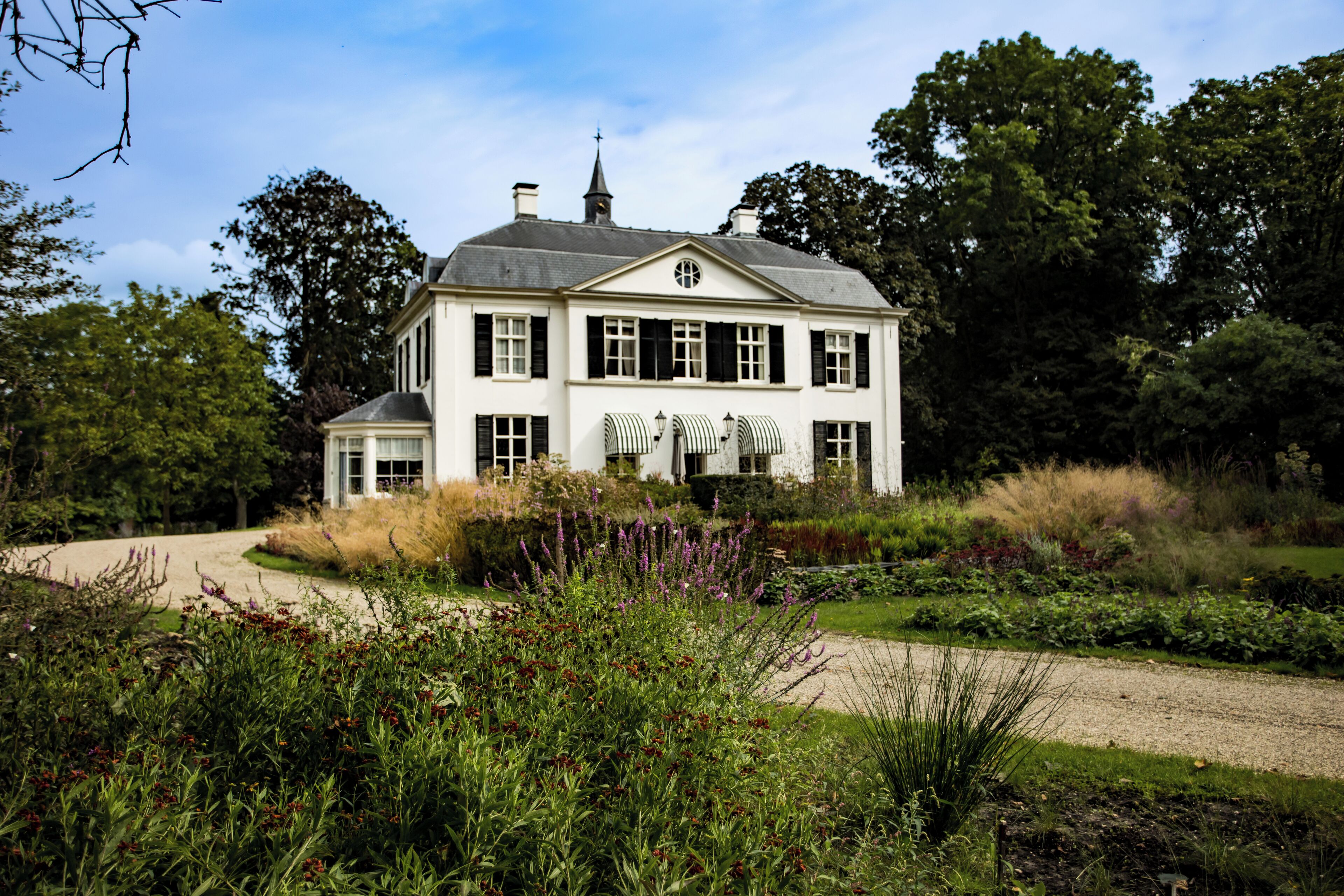 Una casa grande y blanca de dos pisos con fachada simétrica, rodeada de un jardín cuidado con diversas plantas y un camino de grava que lleva a la entrada.