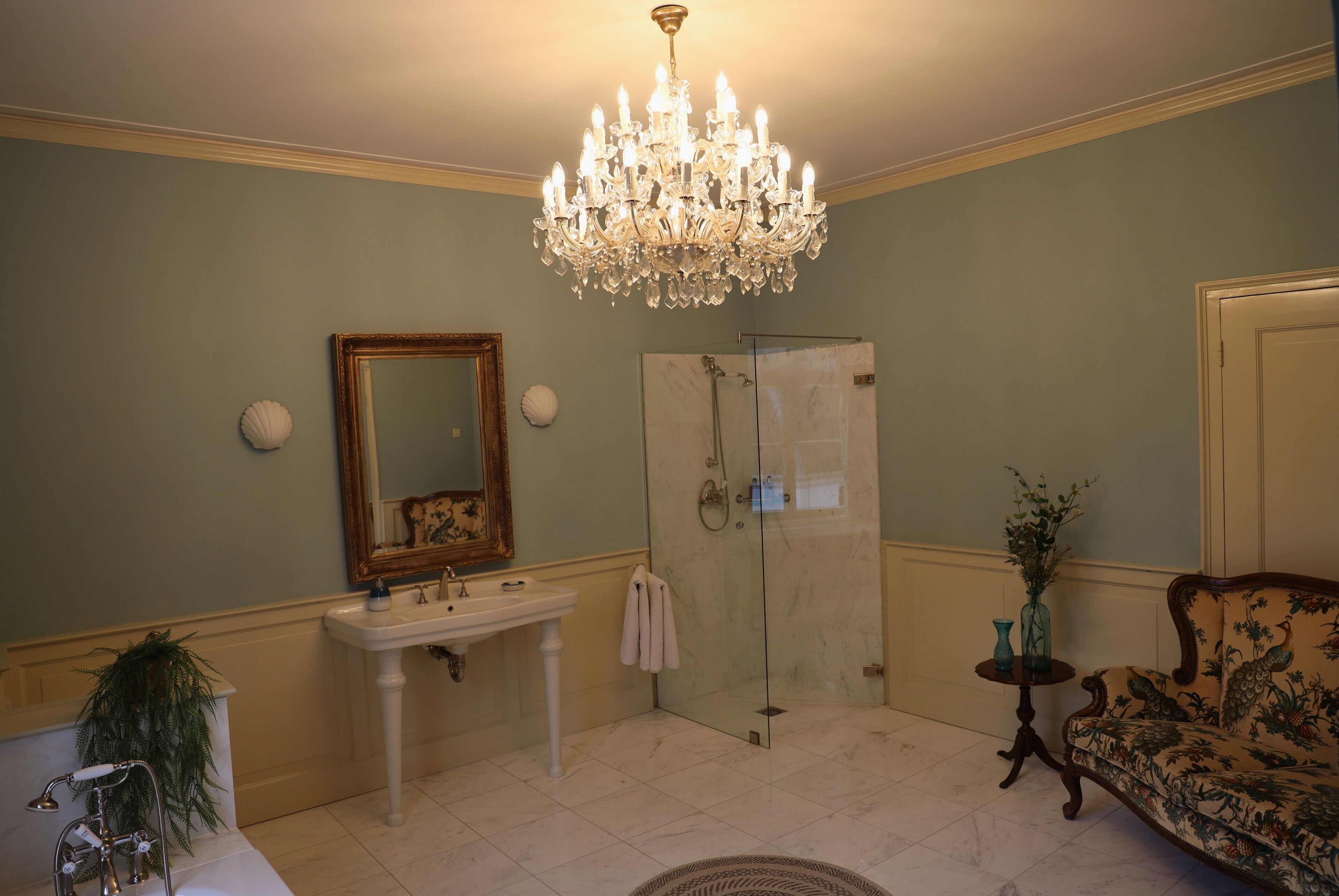 A bathroom with a chandelier, a sink, a mirror, a shower, and a floral sofa.