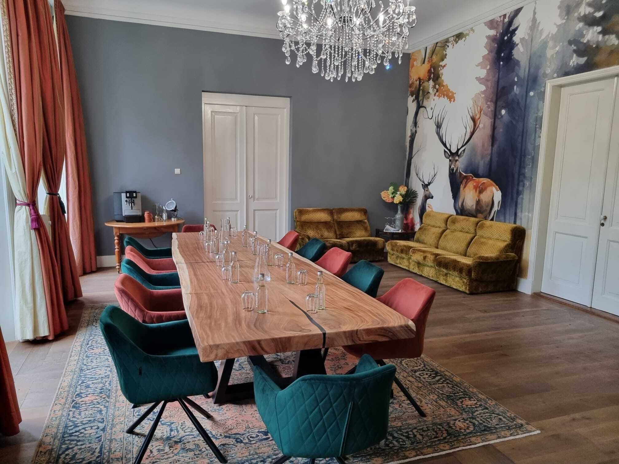 A dining room with a wooden table, colorful chairs, a chandelier, and a painting of deer on the wall.