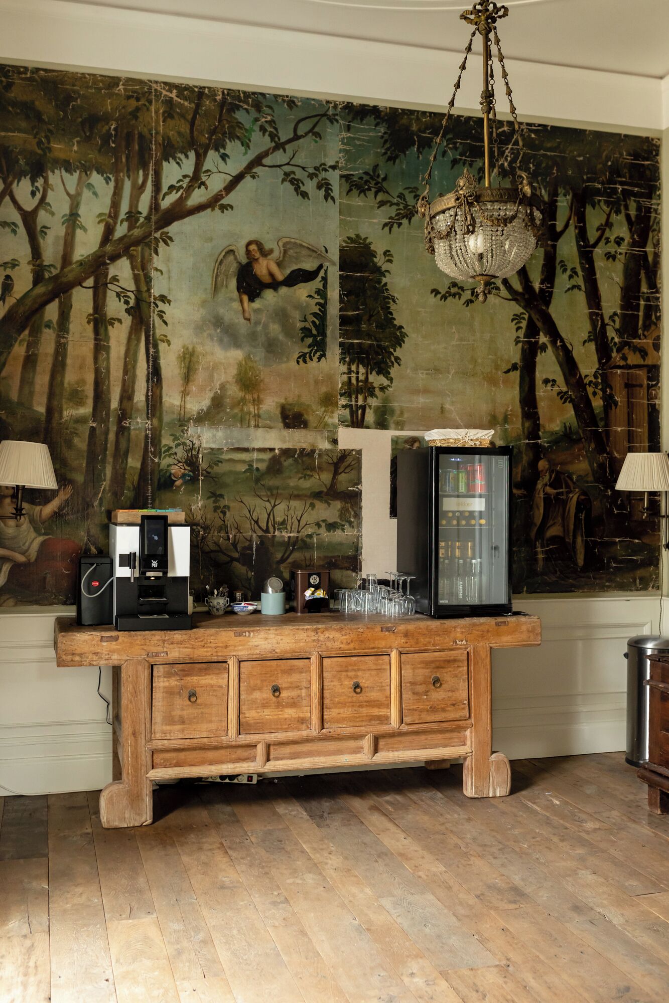 Un coin bar en bois équipé d’une machine à café, d’un réfrigérateur et d’un tableau décoratif représentant un paysage forestier.