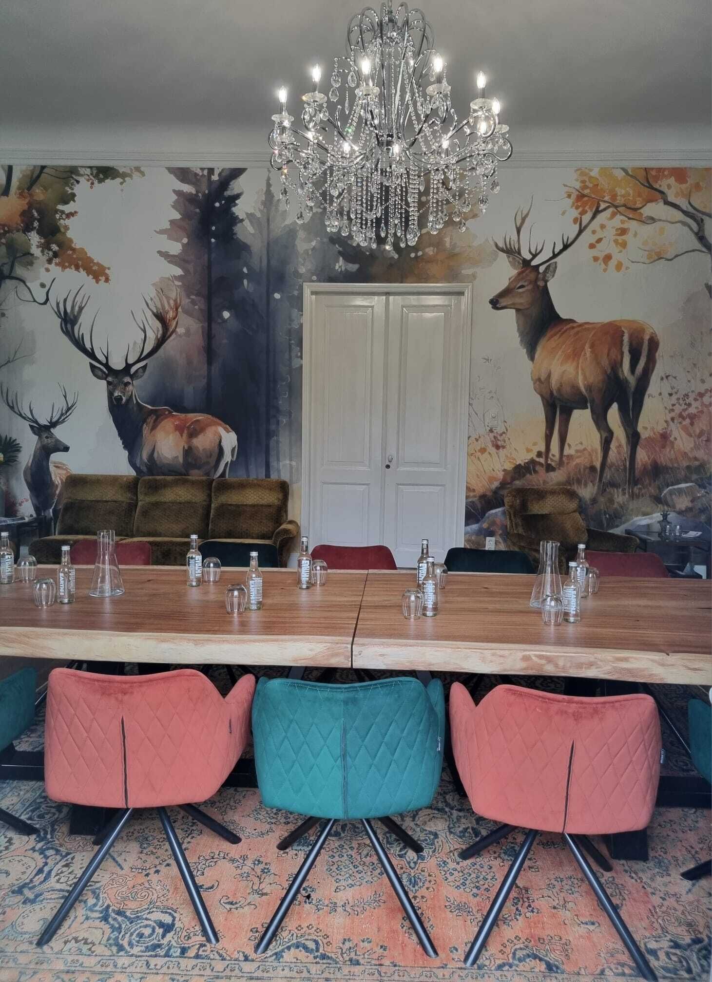 Un comedor con una larga mesa de madera, sillas coloridas, una araña y un mural con motivos de ciervo en la pared.