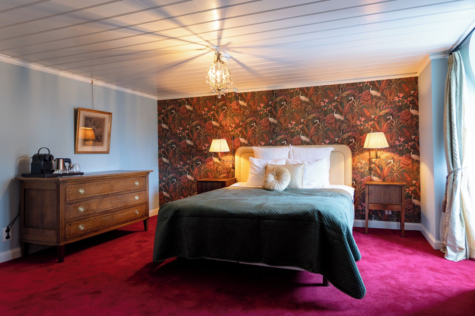 A bedroom with a bed, two bedside tables, a dresser, a chandelier, and a floral wallpaper.