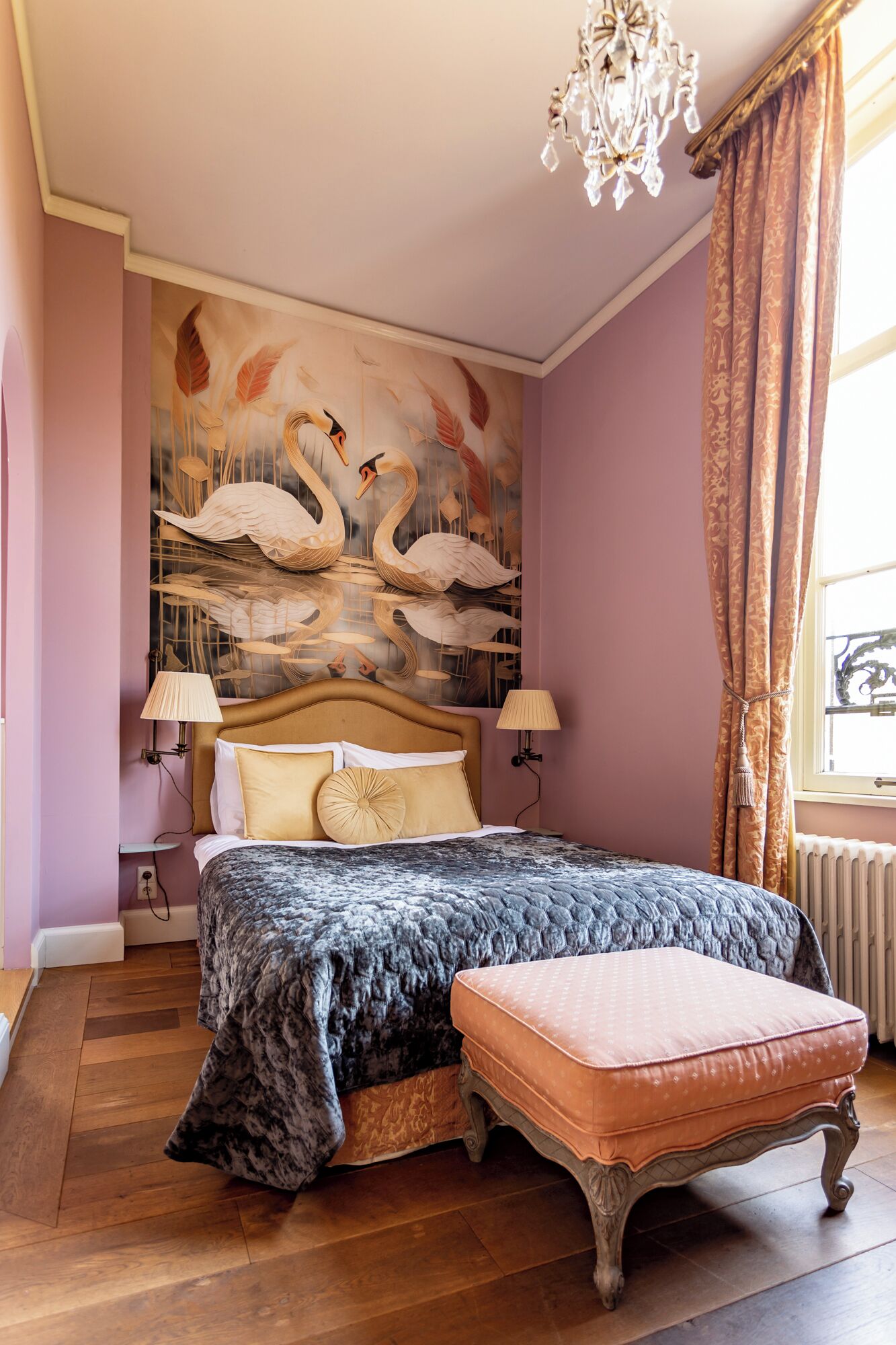 A bedroom with a bed, a nightstand, a wall mural of swans, a chandelier, and a window with curtains.