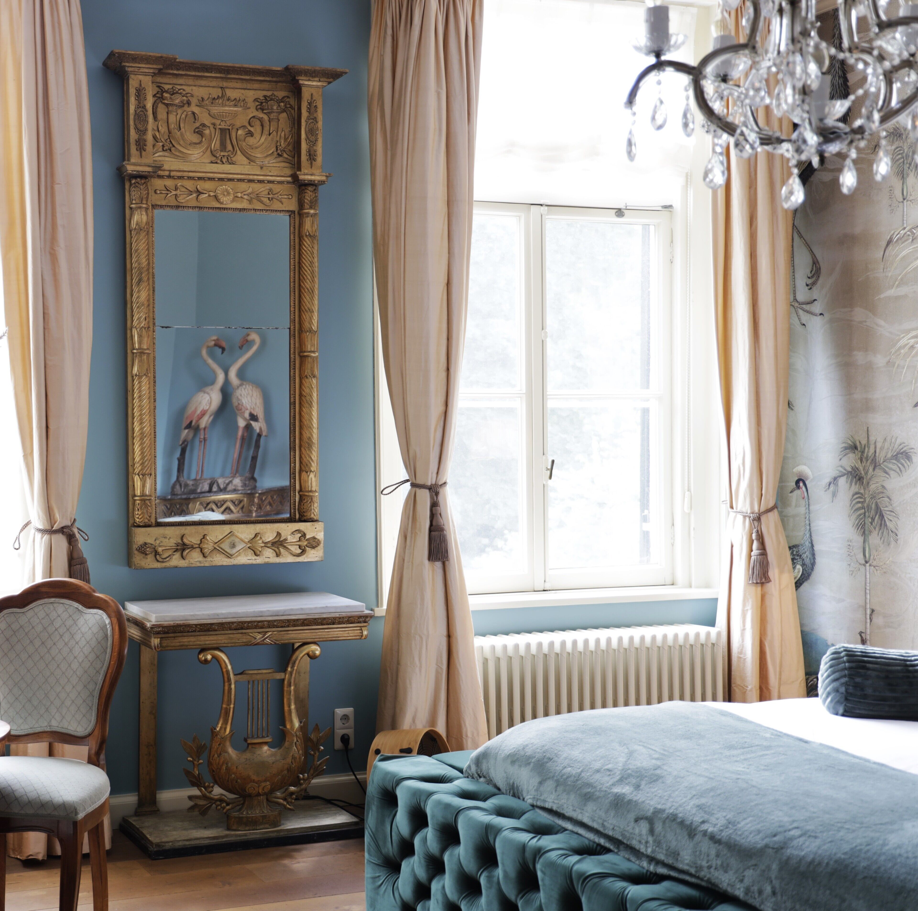 A room with a large mirror, a wooden chair, a bed with a blue upholstered headboard, and a radiator.