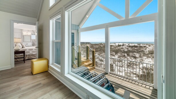 Quarto com grande janela com vista para uma praia, piso de madeira e um pequeno espaço de estar.