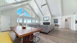 Uma sala de estar espaçosa com pé direito alto, grandes janelas, uma mesa de jantar de madeira e um sofá cinza.