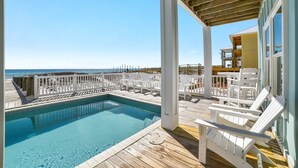 A wooden deck with a pool, white chairs, and a view of the beach.