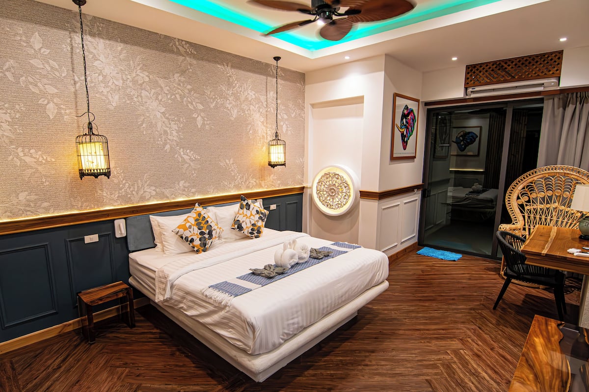 A hotel room with a bed, bedside lamps, a ceiling fan, a dining area with a table and chairs, and a decorative wall piece.