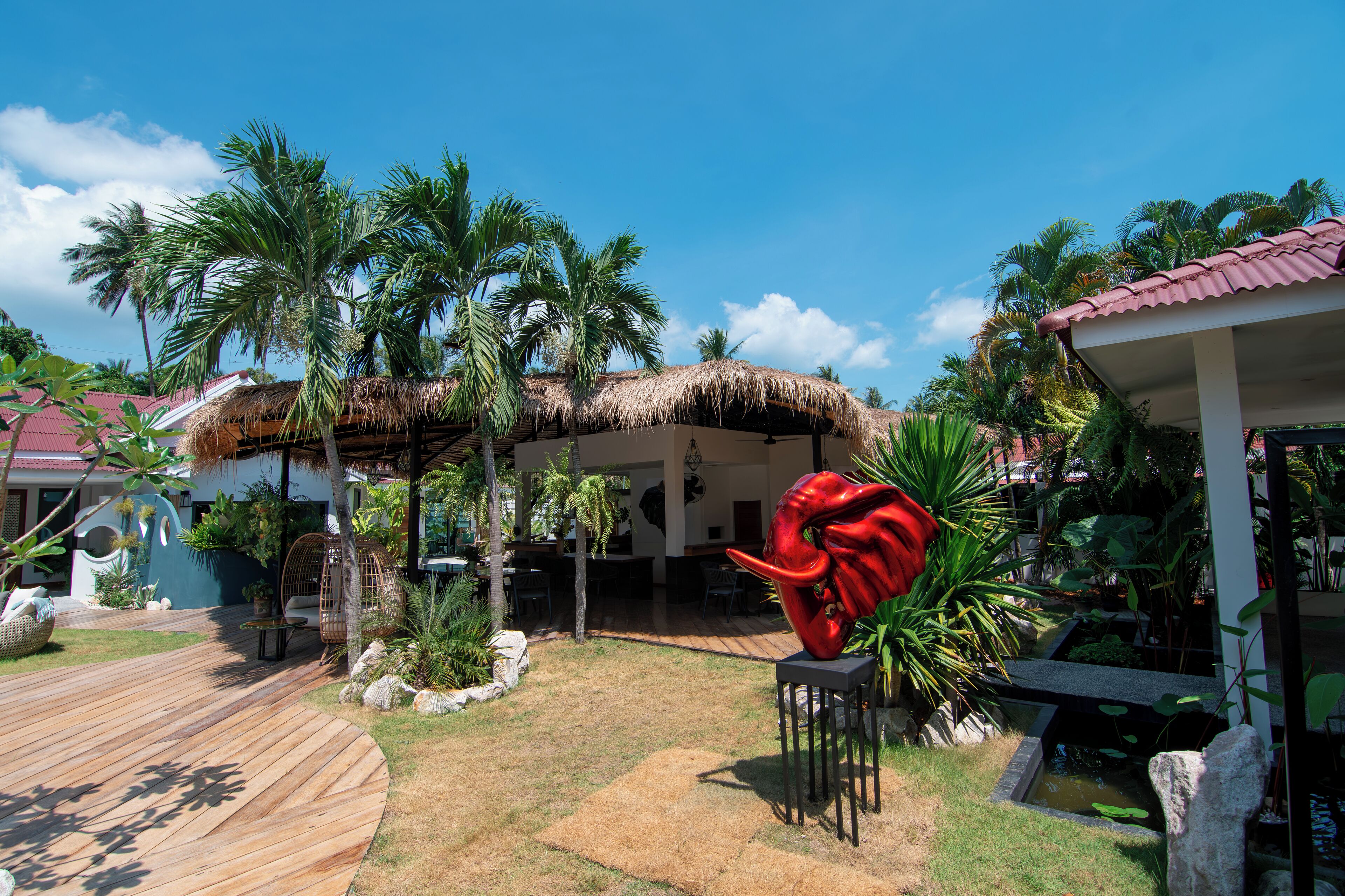 A tropical resort with thatched-roof structures, palm trees, and a red sculpture in the garden.