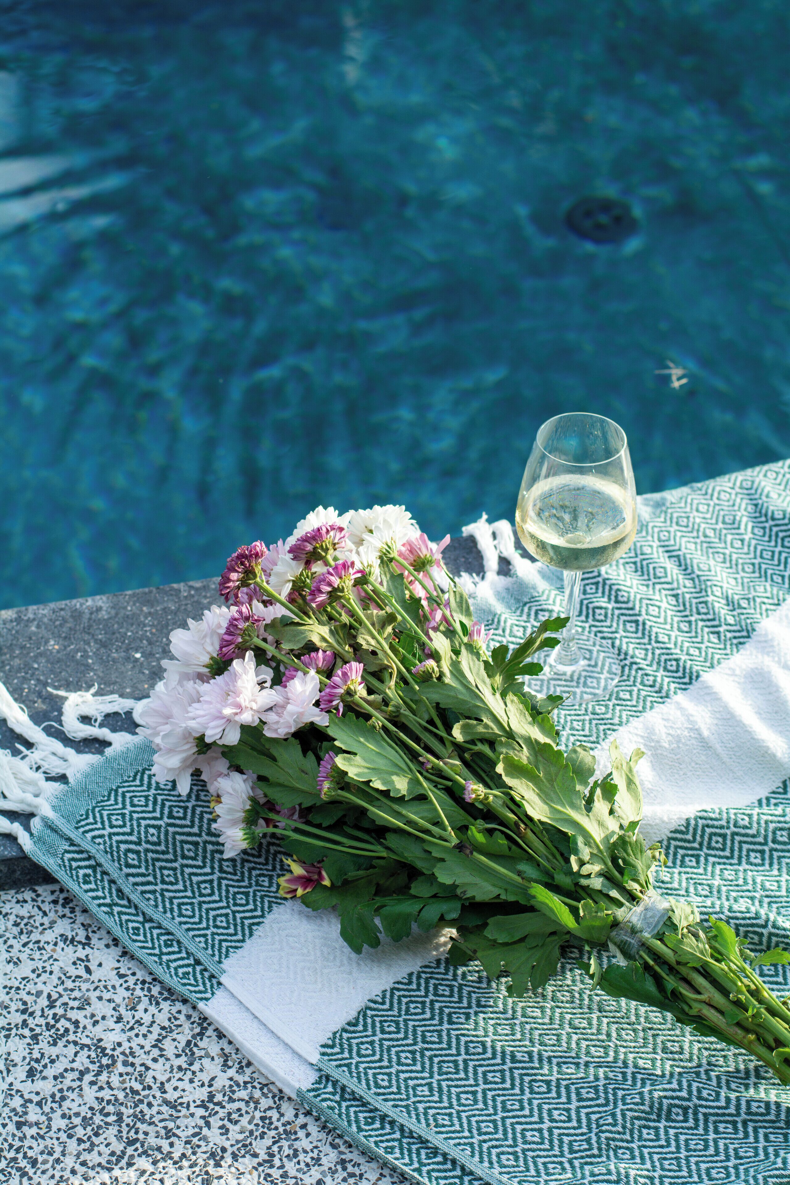 Une scène de bord de piscine agrémentée d’un bouquet de fleurs et d’un verre de vin blanc posé sur une serviette à motifs.