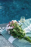 Une scène de bord de piscine agrémentée d’un bouquet de fleurs et d’un verre de vin blanc posé sur une serviette à motifs.