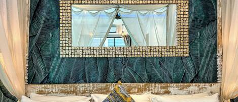 A bedroom with a bed, white pillows, a patterned throw pillow, and a mirror with a mosaic frame.