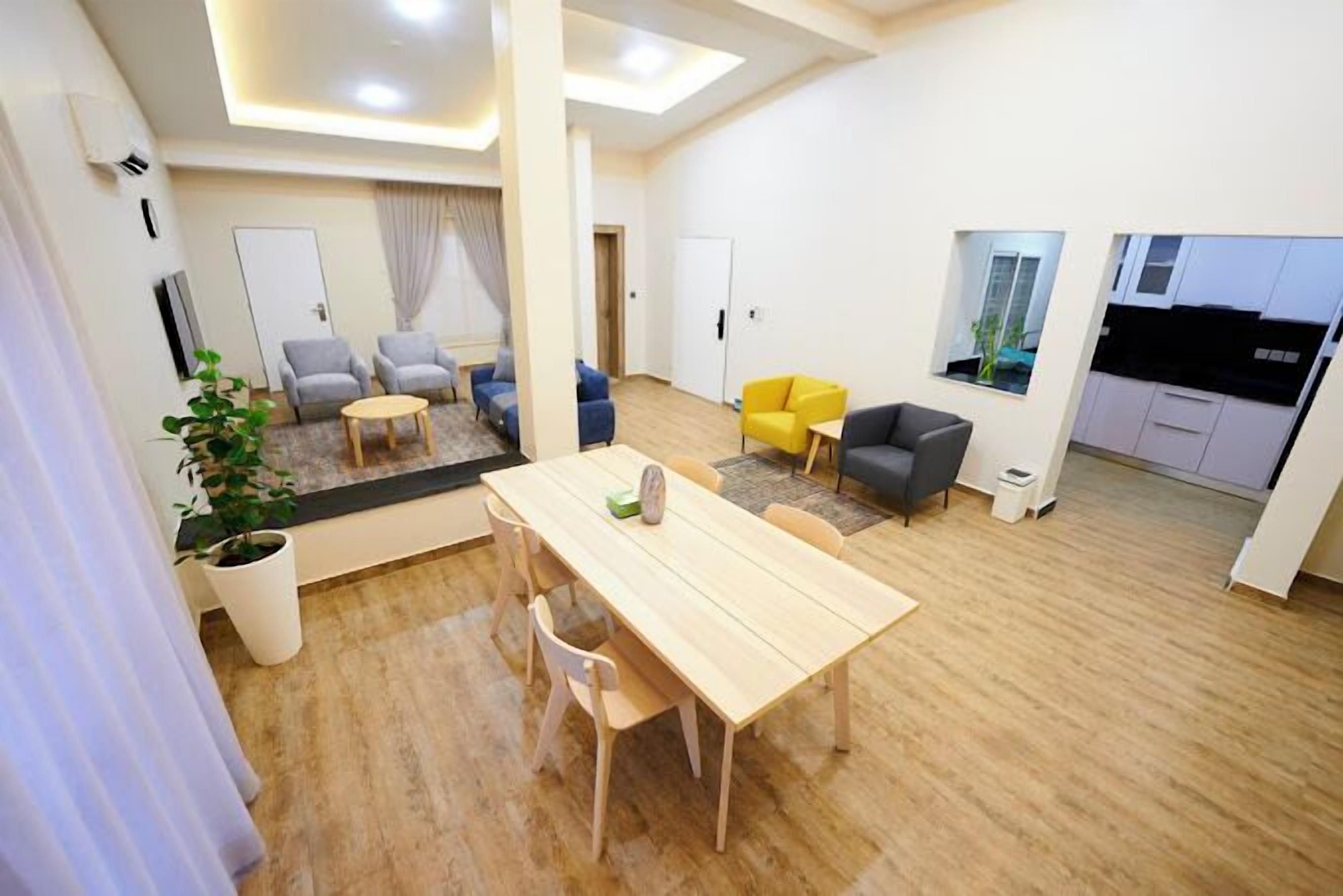Un salon moderne avec une table à manger en bois, des canapés et un coin cuisine visible à travers une cloison en verre.