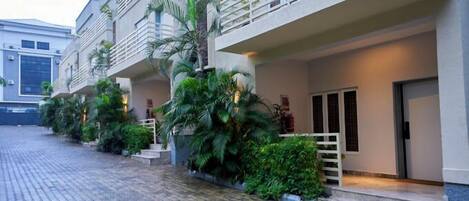 A modern building with a paved courtyard, balconies with railings, and lush greenery.