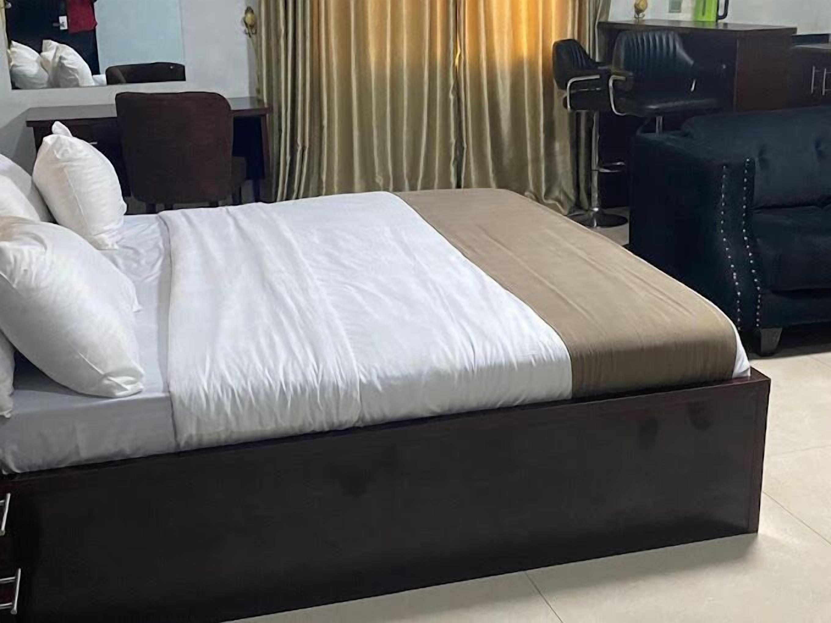 A neatly made bed with white and beige bedding, a dark headboard, and a desk with a chair in the background.