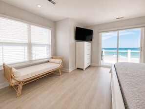 A bedroom with a bed, a TV, a window with blinds, a white wardrobe, and a view of the beach.