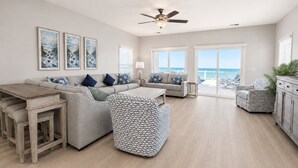 A spacious living room with a large sofa, a dining table, and a patterned ottoman. There are two windows with a view of the ocean. - Oceanfront 7 BR Beach House with a Private Pool! Sandy Days (Surfside Beach)