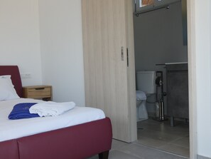 Ein Hotelzimmer mit Bett, Nachttisch aus Holz, Kleiderschrank und einem durch eine offene Tür erreichbaren Badezimmer.
