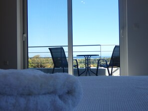 Un balcon avec une table et des chaises, une vue sur la mer et un lit blanc recouvert d’une couverture moelleuse.
