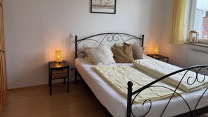 Ein ordentlich bezogenes Bett mit weißen Leinentüchern, zwei Nachttische mit Lampen, ein gerahmtes Bild an der Wand und ein Fenster mit Vorhängen.