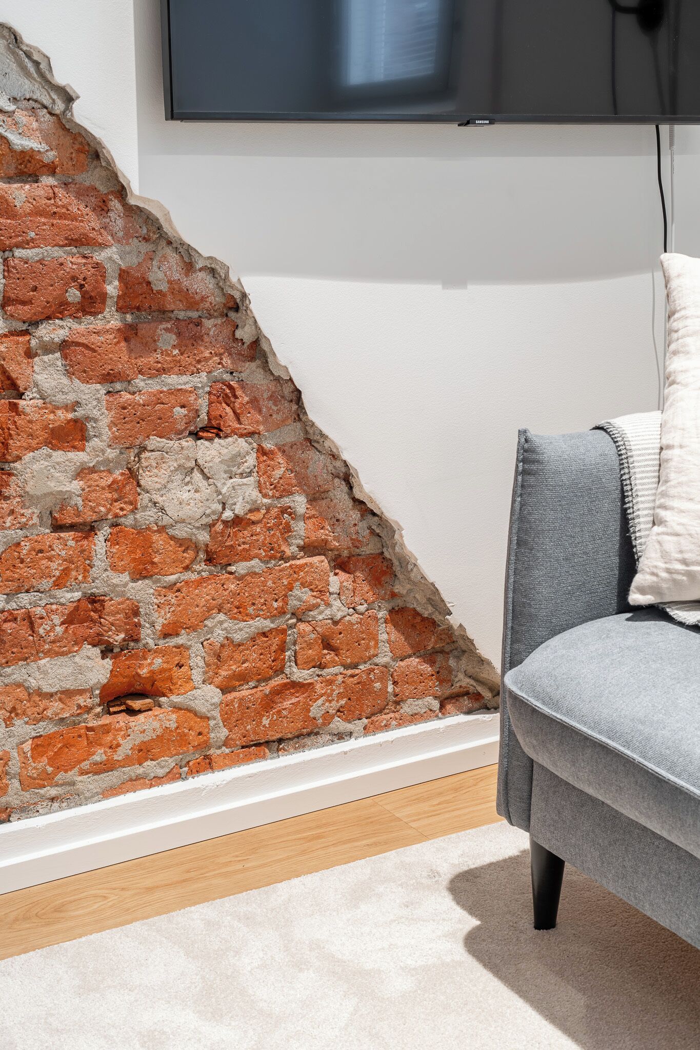 A room with a brick wall, a flat-screen TV mounted on the wall, and a grey sofa with a white cushion.