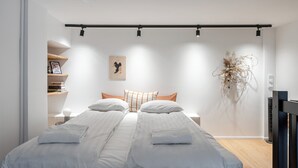 A modern bedroom with a large bed, a small bedside table, a wall-mounted shelf, and a decorative plant.