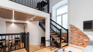 A spacious living area with a wooden floor, a black railing, a staircase with a brick wall, and a flat-screen TV mounted on the wall.