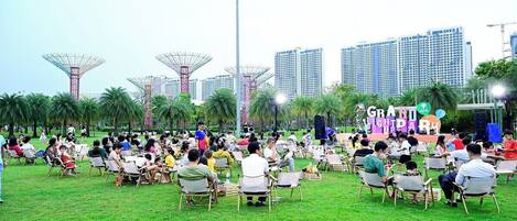 An outdoor event with people seated on chairs in a grassy area, with buildings and a stage in the background.