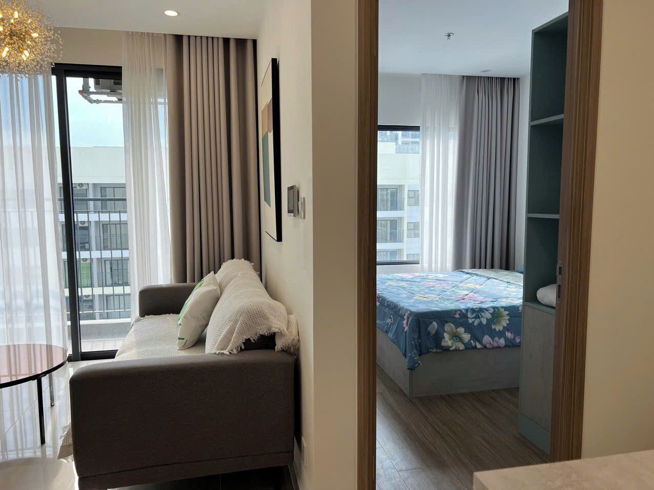 A modern hotel room with a sofa, a bed with a floral bedspread, a small table, and a view of the city through large windows.
