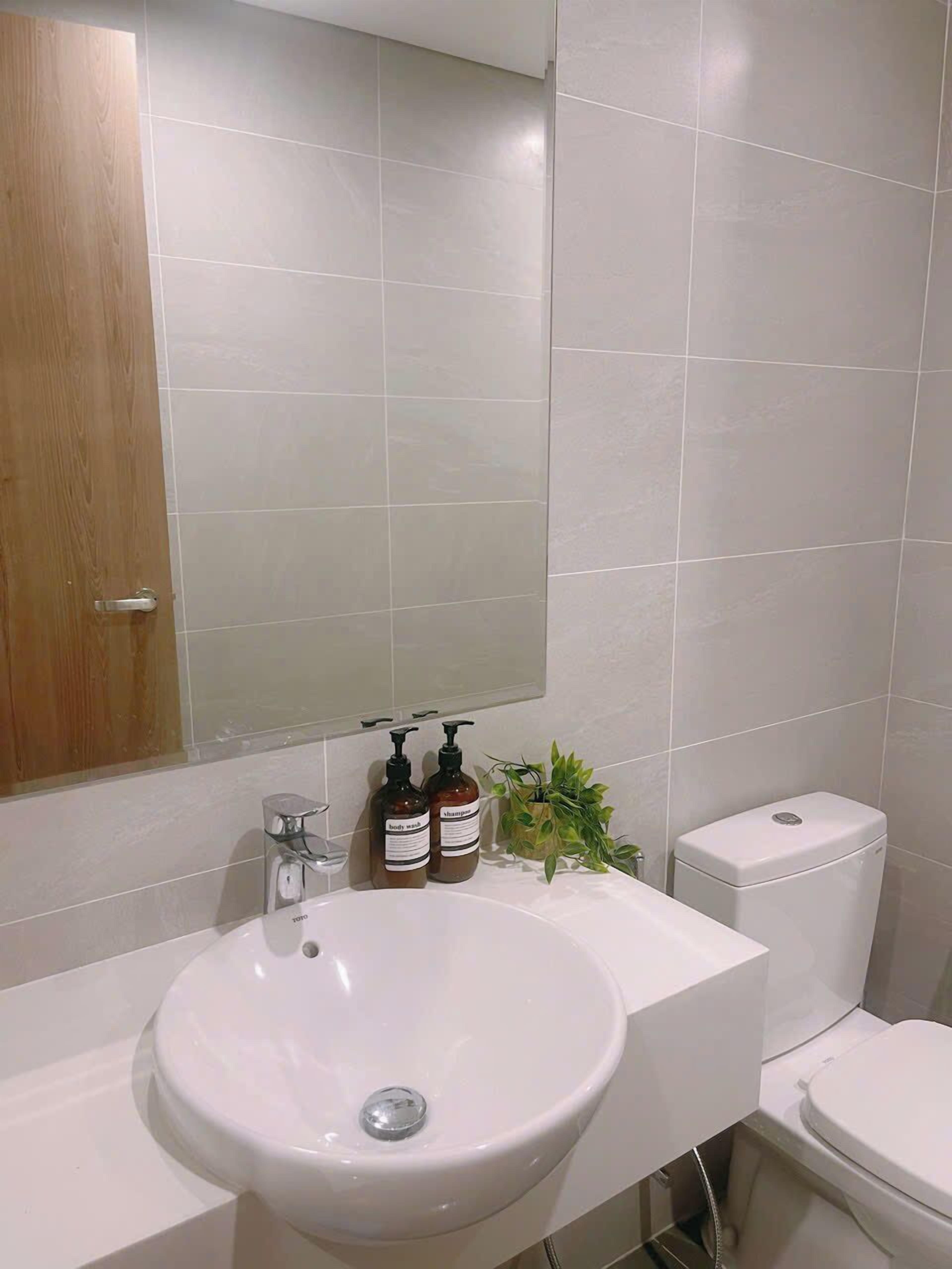 Une salle de bain avec un lavabo blanc, un miroir au-dessus et des toilettes. On y trouve deux bouteilles de shampoing et d’après-shampoing posées sur le lavabo.