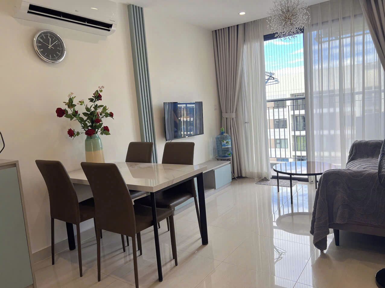 A modern hotel room with a dining area, a sofa, a TV, and a large window with curtains.