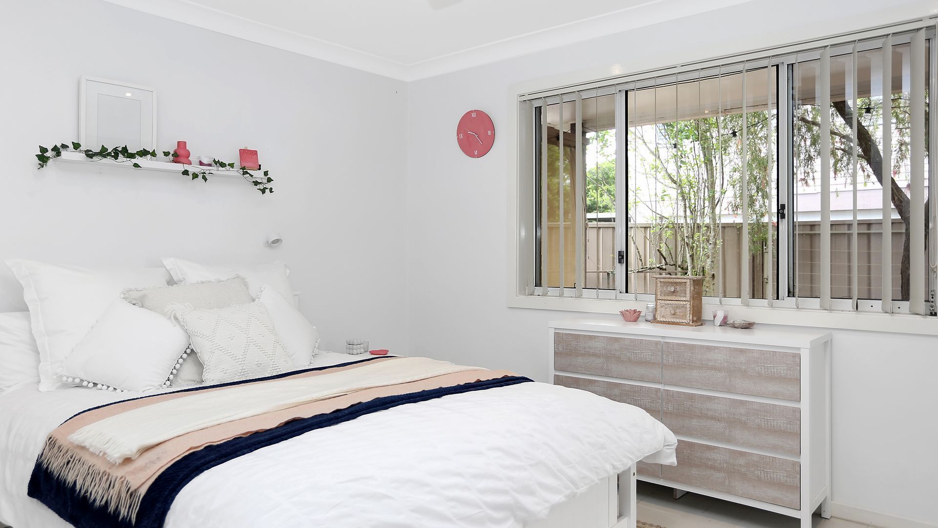 A bedroom with a bed, white walls, a red clock, a window with a view of trees, and a shelf with decorative items.
