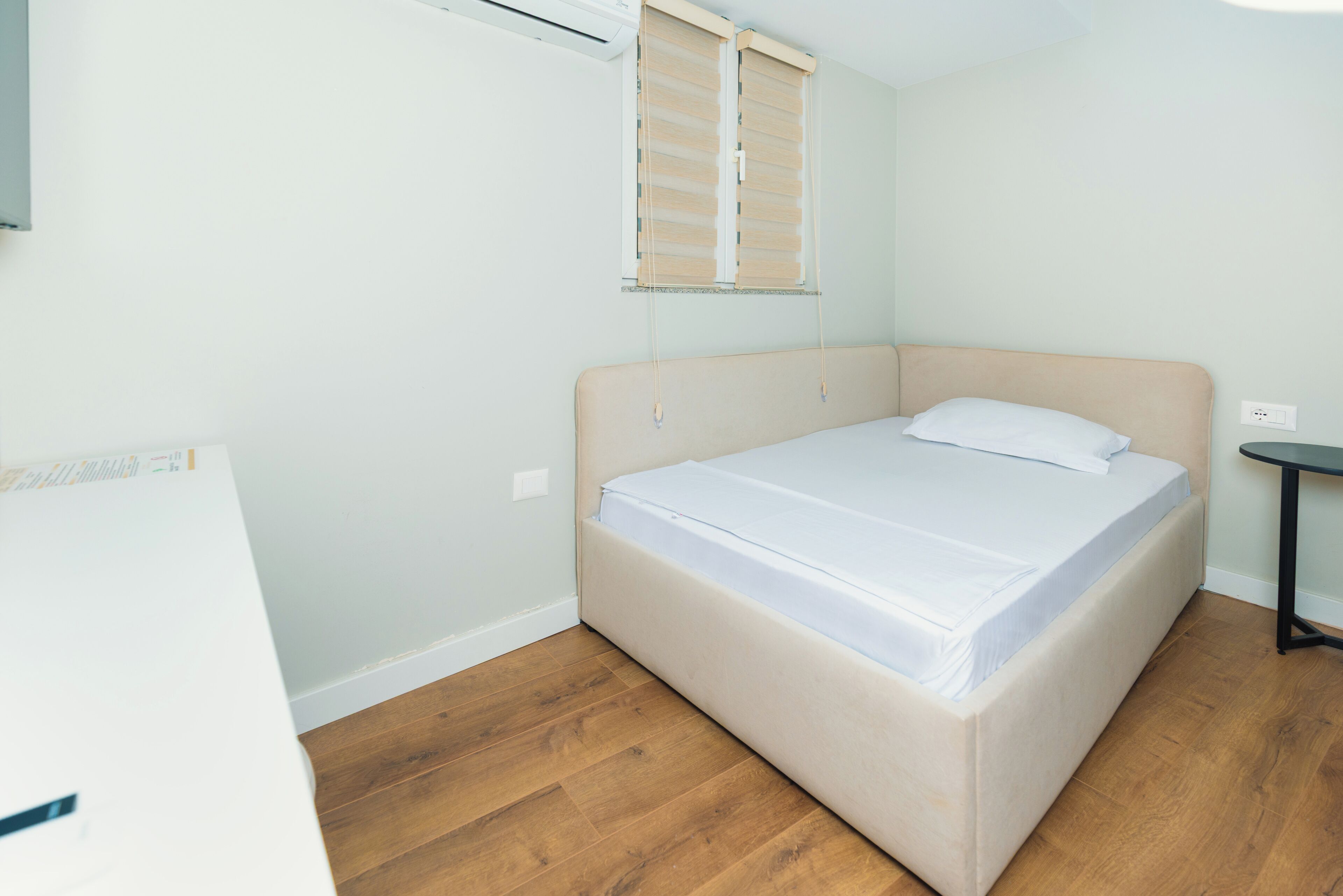 A single bed with a headboard, a bedside table, a window with blinds, and a wall-mounted air conditioner.