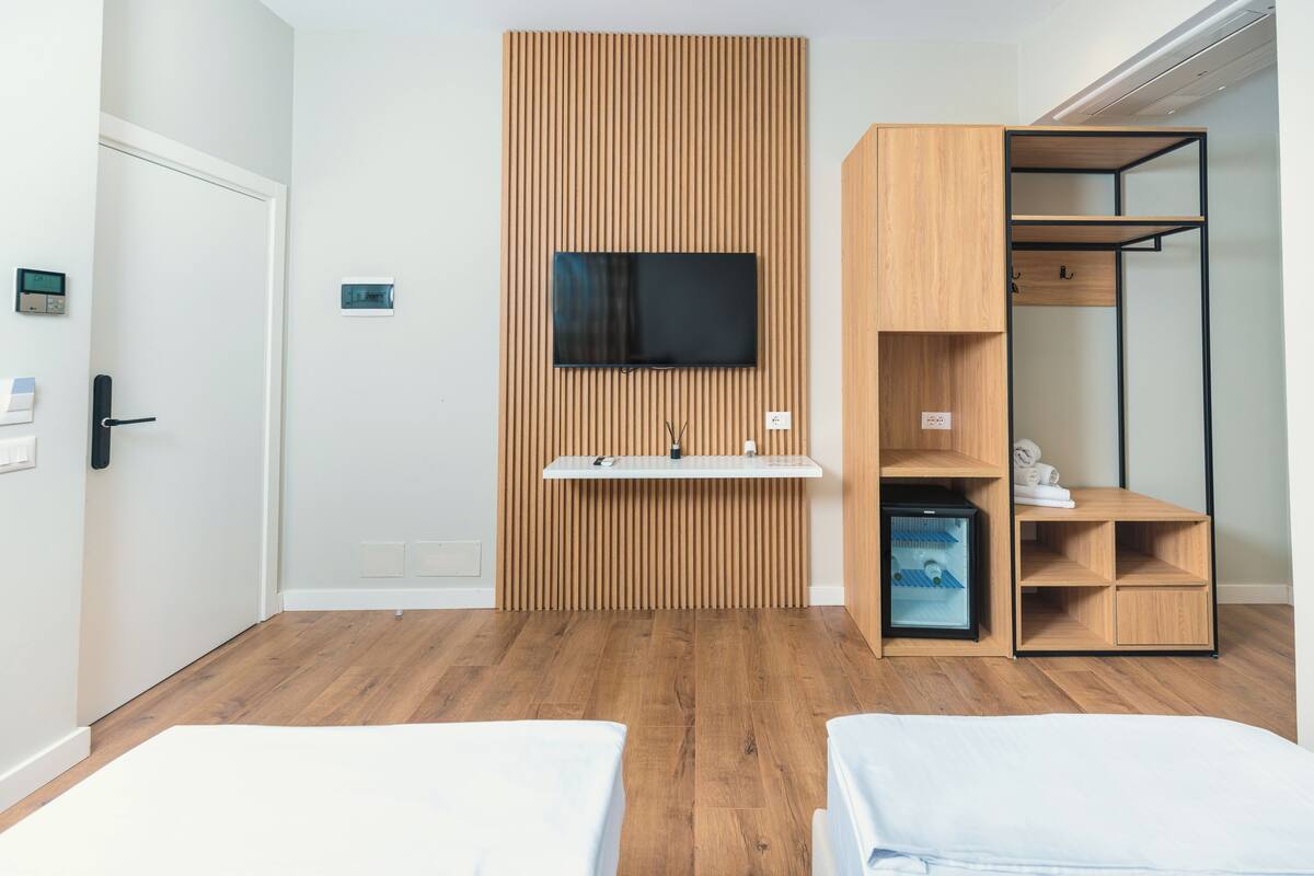 A modern room with a TV mounted on a wooden panel wall, a small shelf, a wardrobe with open shelves, and two beds.