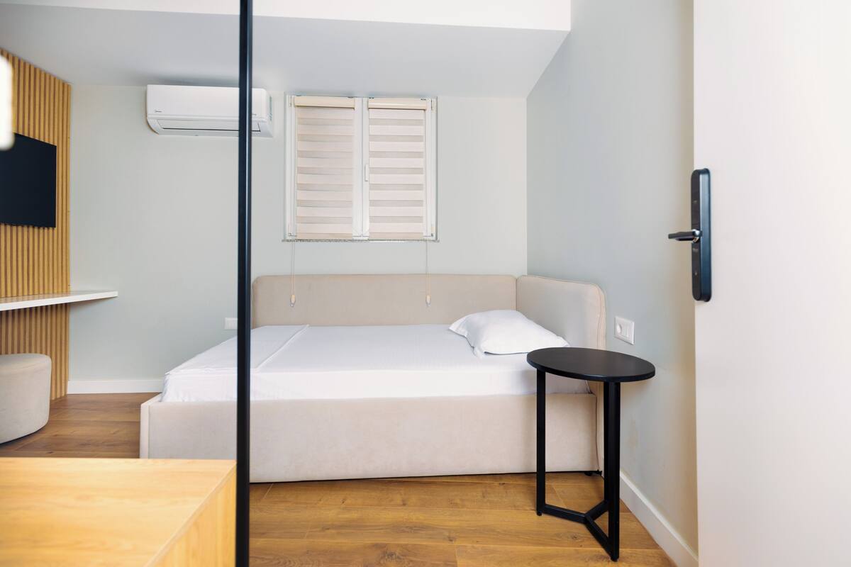 A bedroom with a bed, a small table, a wall-mounted air conditioner, and a window with shutters.