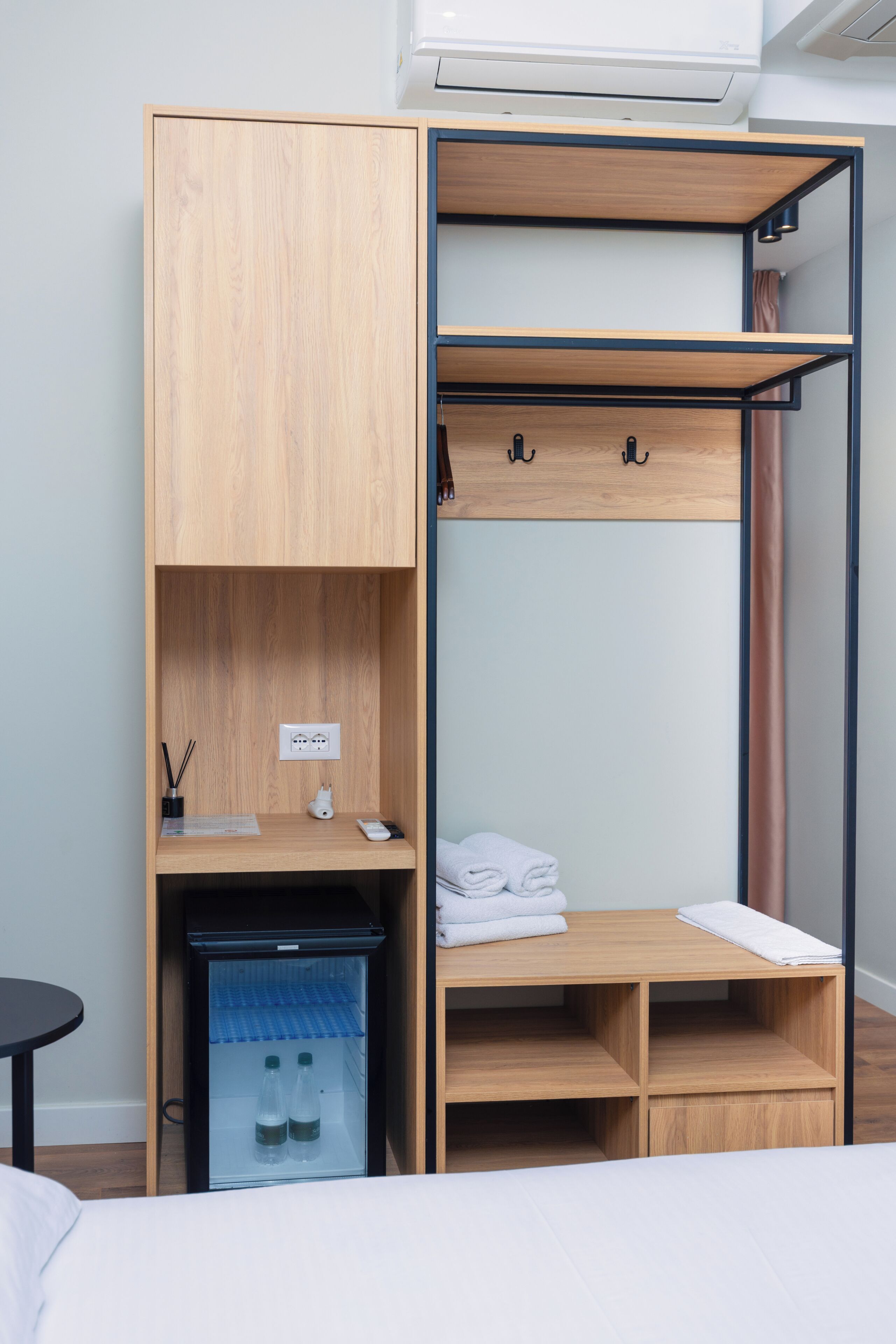 A compact room with a bed, a wardrobe with a built-in desk, a small fridge, and a shelf with towels.