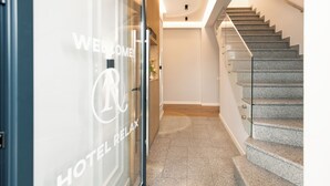 Interior de hotel con escalera, mostrador de recepción y puerta de vidrio con letreros que dicen 'WELCOME' y 'HOTEL RELAX'.