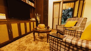A cozy living room with a plaid sofa, a coffee table with decorative items, a bookshelf, and a large flat-screen TV. - Angie's Gardenia a homestay for a comfortable experience  (Candolim)