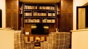 A living room with two plaid armchairs, a coffee table, and a bookshelf filled with books. - Angie's Gardenia a homestay for a comfortable experience  (Candolim)