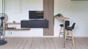 A modern living room with a flat-screen TV, a wooden slat wall, a round clock, a small table with a plant, and two black chairs.