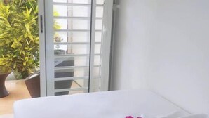 A neatly made bed with white linens and a pink flower, a sliding glass door leading to a balcony with plants, and a white wall.