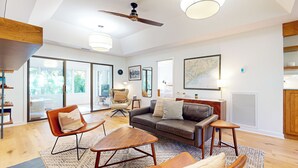 Amplia sala de estar con un sofá moderno, dos sillones, una mesa de centro y un ventilador de techo. Amplias ventanas ofrecen luz natural y vistas al exterior.