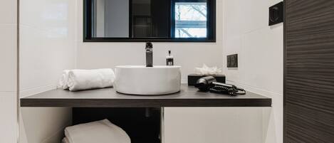 A modern bathroom with a white sink, dark countertop, and a mirror above. There are towels and a toilet paper roll visible.