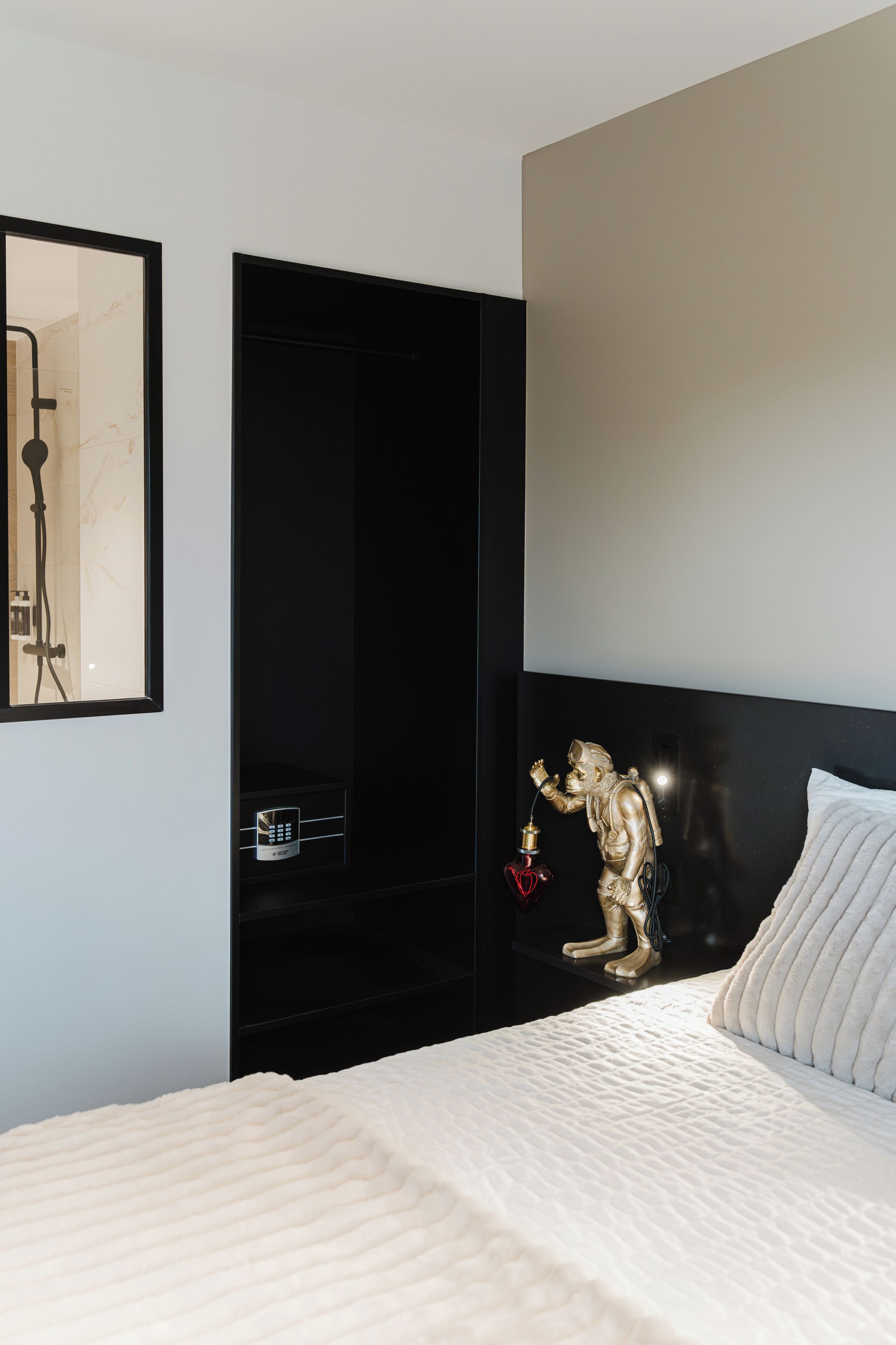 A modern hotel room with a bed, a black wardrobe, a gold statue, and a wall-mounted control panel.