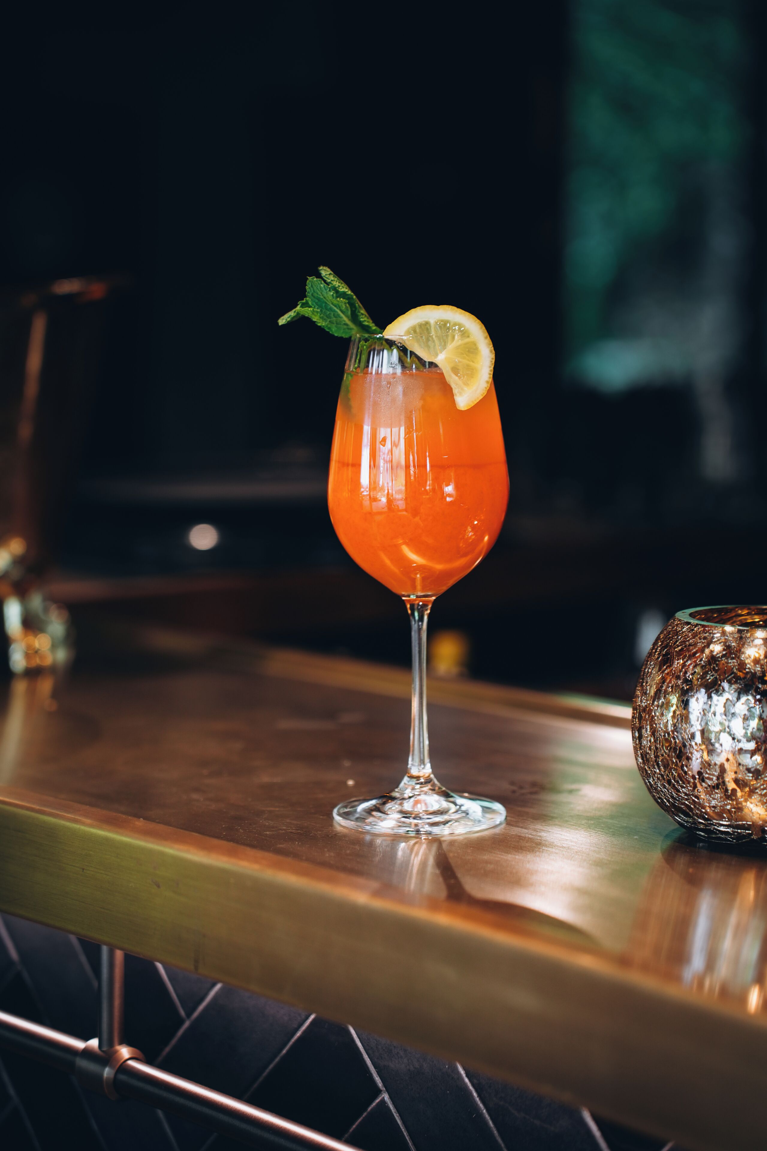 A bar setting with a glass of orange-colored drink garnished with a lemon slice and mint, placed on a wooden bar counter.