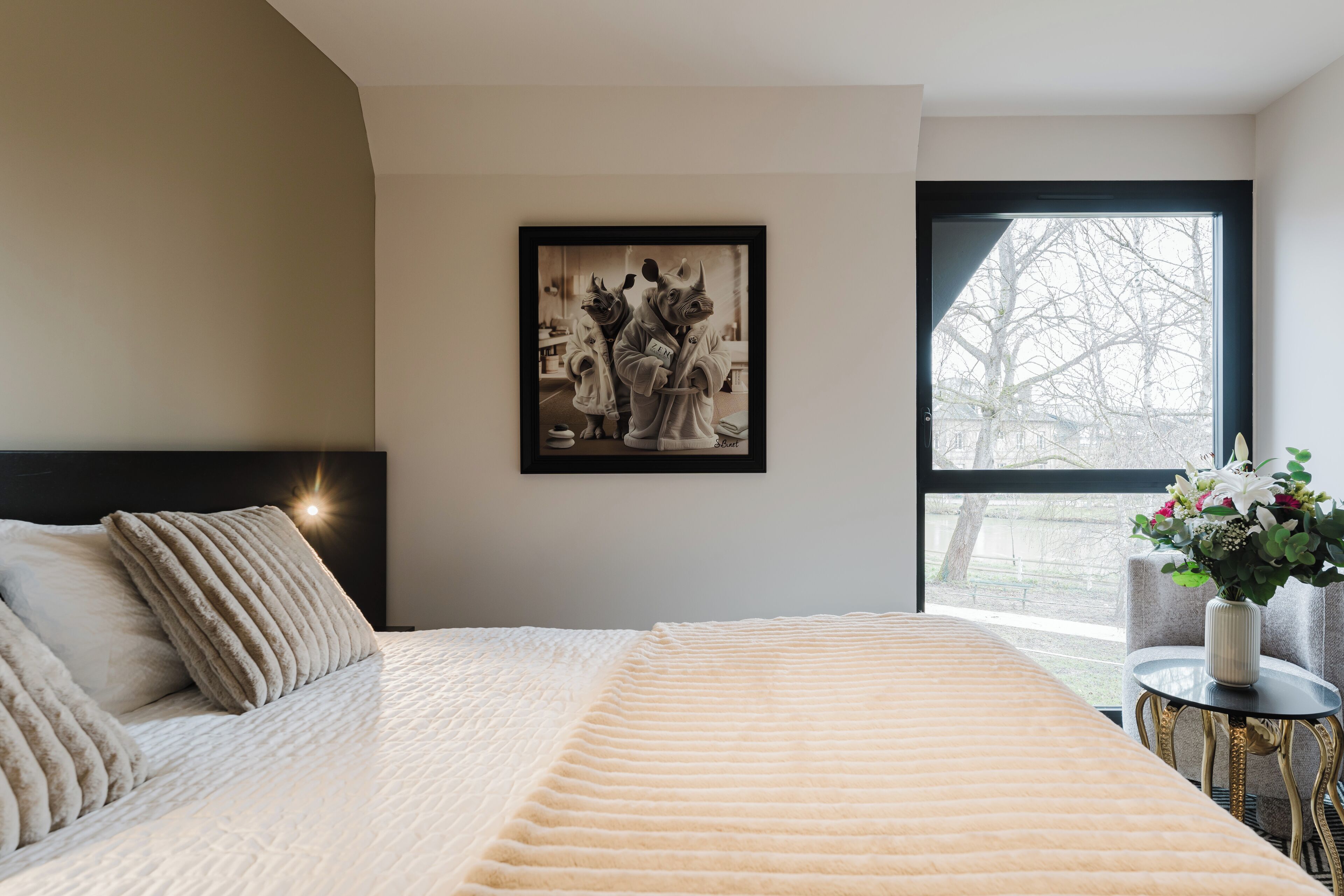 A bedroom with a large bed, a framed artwork on the wall, and a window with a view of trees.