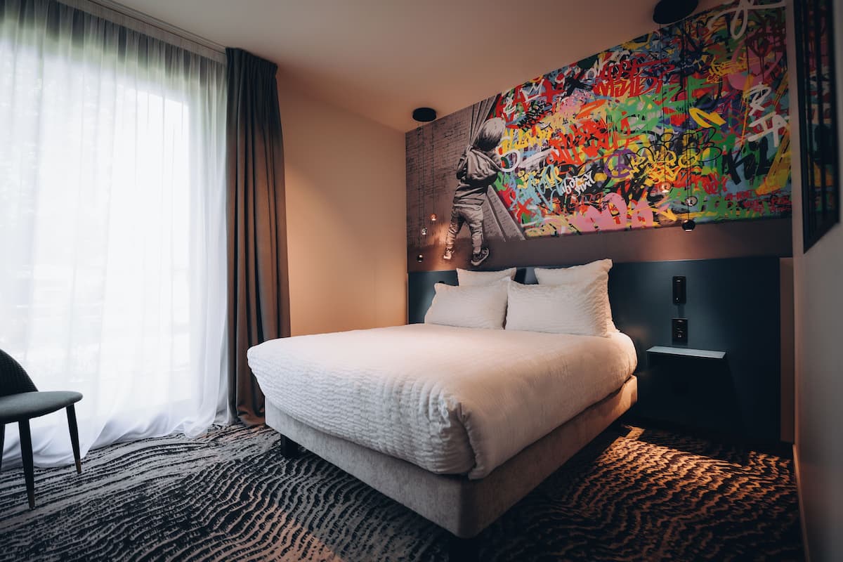 Ein Hotelzimmer mit Bett, Sessel, großem abstrakten Gemälde an der Wand und einem gemusterten Teppich.